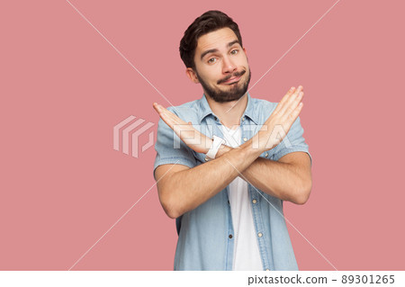 Portrait of serious handsome bearded young man in blue casual style shirt standing with X sign hands and looking at camera. indoor studio shot, isolated on pink background. 89301265