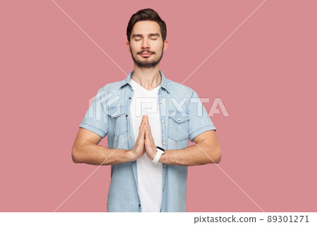 Portrait of calm relax handsome bearded young man in blue casual style shirt standing with palm hands, closed eyes and doing yoga meditation exercise. indoor studio shot, isolated on pink background. Portrait of calm relax handsome bearded young man in blue casual style shirt standing with palm hands, closed eyes and doing yoga meditation exercise. indoor studio shot, isolated on pink background. 89301271