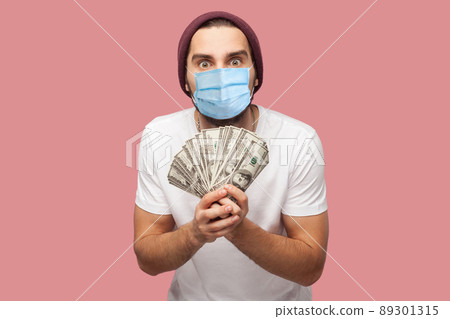 Money. Portrait of suprised young hipster man with surgical medical mask in white shirt and casual hat, stand, holding fan of dollars with big eyes. Indoor, isolated, studio shot, pink background Money. Portrait of suprised young hipster man with surgical medical mask in white shirt and casual hat, stand, holding fan of dollars with big eyes. Indoor, isolated, studio shot, pink background 89301315