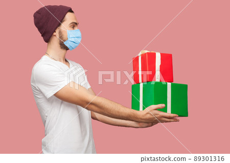 Side view portrait of friendly young hipster man with surgical medical mask in white shirt and casual hat standing, giving two present box and smile. Indoor, isolated, studio shot, pink background Side view portrait of friendly young hipster man with surgical medical mask in white shirt and casual hat standing, giving two present box and smile. Indoor, isolated, studio shot, pink background 89301316