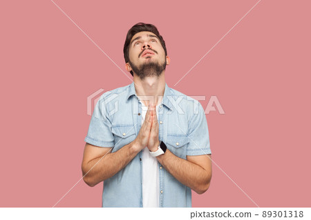 My God please help me. Portrait of hopeful sad handsome bearded young man in blue casual style shirt standing looking up and praying with worry face. indoor studio shot, isolated on pink background. My God please help me. Portrait of hopeful sad handsome bearded young man in blue casual style shirt standing looking up and praying with worry face. indoor studio shot, isolated on pink background. 89301318