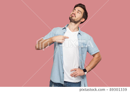 This is me. Portrait of proud haughty handsome bearded young man in blue casual style shirt standing, looking away and pointing himself. indoor studio shot, isolated on pink background. 89301339