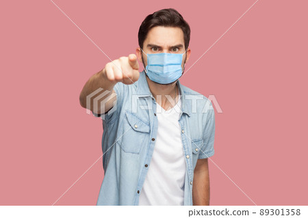 Hey you. Portrait of serious man with surgical medical mask in blue casual style shirt standing pointing and looking at camera with angry face. indoor studio shot, isolated on pink background. Hey you. Portrait of serious man with surgical medical mask in blue casual style shirt standing pointing and looking at camera with angry face. indoor studio shot, isolated on pink background. 89301358