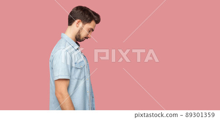 Profile side view portrait of sad alone or confused handsome bearded young man in blue casual style shirt standing, holding his head down and thinking. indoor studio shot, isolated on pink background. Profile side view portrait of sad alone or confused handsome bearded young man in blue casual style shirt standing, holding his head down and thinking. indoor studio shot, isolated on pink background. 89301359