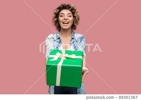 Portrait of happy beautiful young woman in blue shirt with curlty hairstyle standing and giving you green gift box with toothy smile, looking at camera. Studio shot, pink background, isolated, indoor 89301367