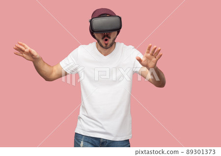Portrait of suprised bearded young hipster man in white shirt and casual hat standing, wearing vr and watching video on simulator with raised arms. Indoor, isolated, studio shot, pink background 89301373