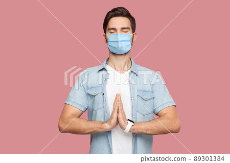 Portrait of calm relax young man with surgical medical mask in blue shirt standing with palm hands, closed eyes and doing yoga meditation exercise. indoor studio shot, isolated on pink background. 89301384