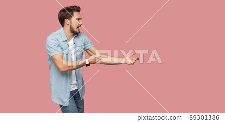 Profile side view portrait of serious screaming bearded young man in blue casual shirt standing in attack or pulling hands gesture and looking forward. indoor studio shot, isolated on pink background. 89301386