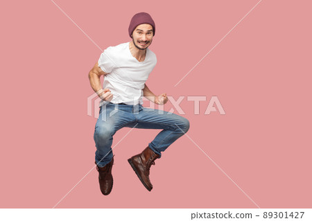 Portrait of happy bearded hipster young man in white shirt and blue jeans with hat jumping, rejoicing and celebrating his victory. indoor studio shot, isolated on pink background copyspace. 89301427