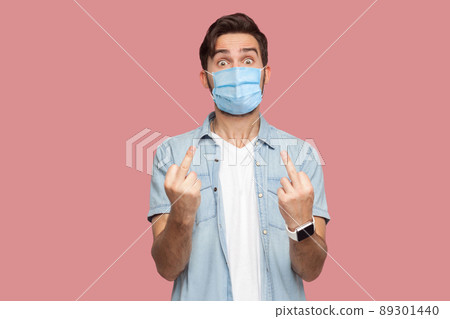 Portrait of angry young man with surgical medical mask in blue shirt standing with middle finger fuck sign hand gesture and looking at camera. indoor studio shot, isolated on pink background. Portrait of angry young man with surgical medical mask in blue shirt standing with middle finger fuck sign hand gesture and looking at camera. indoor studio shot, isolated on pink background. 89301440