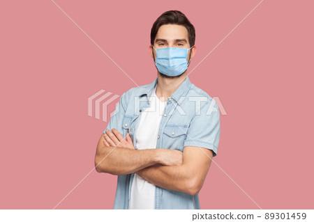 Portrait of handsome young man with surgical medical mask in blue casual style shirt standing, raised arms and looking at camera with smile. indoor studio shot, isolated on pink background. Portrait of handsome young man with surgical medical mask in blue casual style shirt standing, raised arms and looking at camera with smile. indoor studio shot, isolated on pink background. 89301459