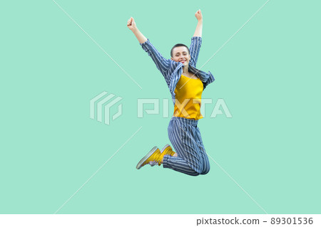 Portrait of successful beautiful short hair young stylish woman in casual striped suit jumping and celebraiting her victory, looking at camera. indoor studio shot isolated on light green background. 89301536