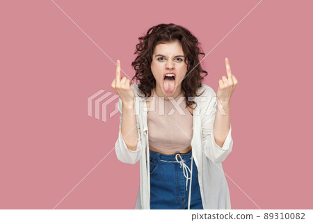 Portrait of angry screaming brunette young woman with curly hairstyle in casual style standing and looking at camera with middle finger fuck sign. indoor studio shot isolated on pink background. 89310082