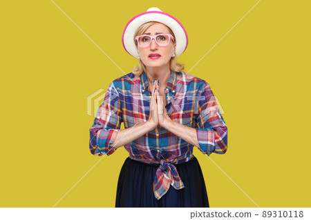 Please forgive me. or give me one more chance, Portrait of hopeful mature woman in casual style with hat and eyeglasses standing with palm hands and looking at camera, begging and asking. studio shot. 89310118