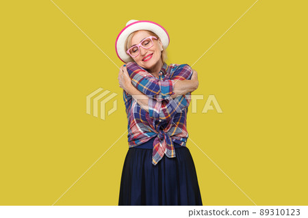 Portrait of peacful happy modern stylish mature woman in casual style with hat, eyeglasses standing, hugging herself with closed eyes and toothy smile. indoor studio shot isolated on yellow background 89310123