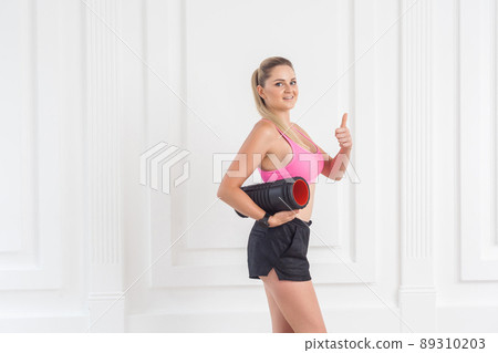 Sporty concept. Satisfied athletic young beautiful blonde girl in pink top and black shorts standing, holding foam roll and showing thumbs up. Studio shot, looking at camera, indoor, isolated Sporty concept. Satisfied athletic young beautiful blonde girl in pink top and black shorts standing, holding foam roll and showing thumbs up. Studio shot, looking at camera, indoor, isolated 89310203