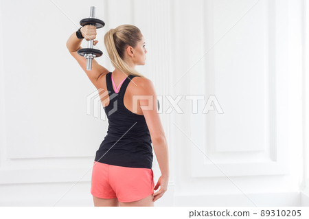 Striving for strong. Backside of young adult motivation athletic bodybuilder woman in pink shorts and black top standing, holding dumbells doing exercise for arms at the gym with perfect figure. Striving for strong. Backside of young adult motivation athletic bodybuilder woman in pink shorts and black top standing, holding dumbells doing exercise for arms at the gym with perfect figure. 89310205