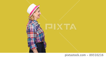 Profile side view portrait of happy successful modern stylish mature woman in casual style with hat standing, looking forward with toothy smile. indoor studio shot isolated on yellow background. Profile side view portrait of happy successful modern stylish mature woman in casual style with hat standing, looking forward with toothy smile. indoor studio shot isolated on yellow background. 89310216