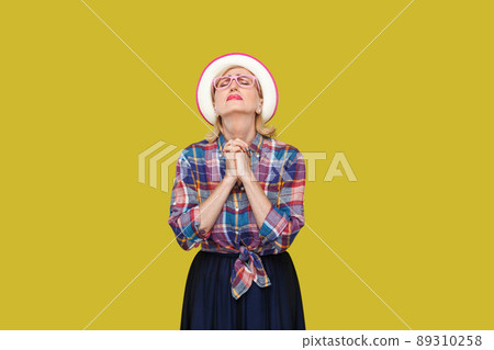 Oh my God please help me. Portrait of hopeful modern stylish mature woman in casual style with hat, eyeglasses standing with closed eyes and praying. indoor studio shot isolated on yellow background. 89310258