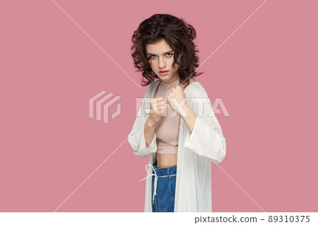 Portrait of serious beautiful brunette young woman with curly hairstyle in casual style standing in boxing pose, looking at camera and ready to attack. indoor studio shot isolated on pink background. Portrait of serious beautiful brunette young woman with curly hairstyle in casual style standing in boxing pose, looking at camera and ready to attack. indoor studio shot isolated on pink background. 89310375