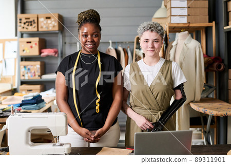 Two happy young intercultural fashion designers standing by workplace equipped with electric sewing-machine in spacious workshop Two happy young intercultural fashion designers standing by workplace equipped with electric sewing-machine in spacious workshop 89311901