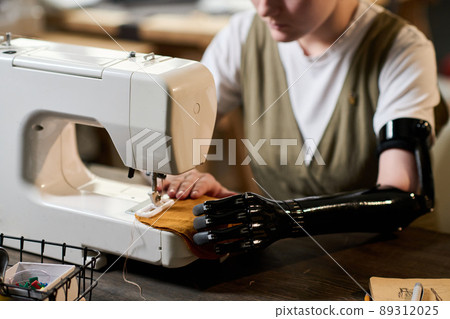 Close-up of young female with myoelectric arm prosthesis sewing clothes by wooden table while creating new items for fashion collection 89312025