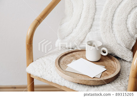 Trendy Scandinavian interior still life. Breakfast concept. Blank greeting card, invitation and cup of coffee on wooden tray. Stationery mock-up. Wooden chair, wool teddy armchair, white cushion. 89315205
