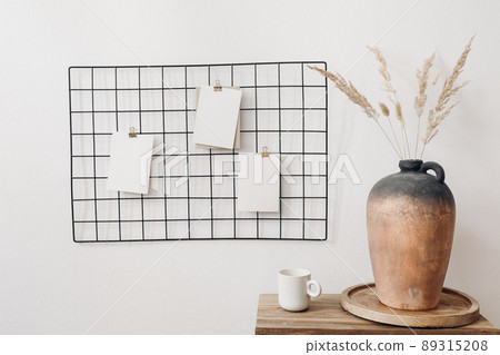 Black metal mesh noticeboard, bulletin board with blank memo cards mockups. Elegant home office interior concept. Vase with dry lagurus grass and cup of coffe on wooden table. White wall background. Black metal mesh noticeboard, bulletin board with blank memo cards mockups. Elegant home office interior concept. Vase with dry lagurus grass and cup of coffe on wooden table. White wall background. 89315208