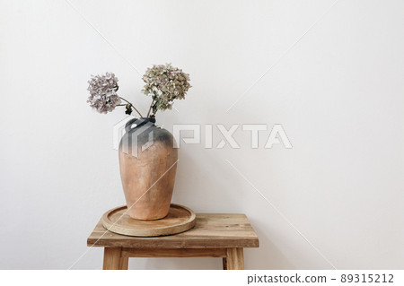 Elegant minimal summer, fall still life photo. Rustic clay vase, pitcher with hydrangea flowers on old wooden stool, console table. White wall background. Empty copy space. Elegant interior, lifestyle Elegant minimal summer, fall still life photo. Rustic clay vase, pitcher with hydrangea flowers on old wooden stool, console table. White wall background. Empty copy space. Elegant interior, lifestyle 89315212
