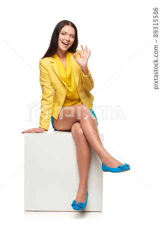 Full length of smiling girl in yellow and blue clothes sitting on white box gesturing OK 89315586