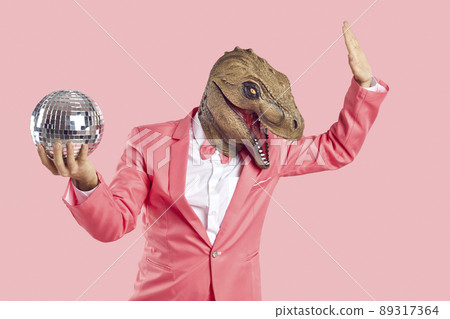 Fashionable man in dinosaur rubber mask with disco ball in his hand on pastel pink background. 89317364