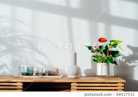Modern minimalist Scandinavian style interior. Candles, ceramic vase and House plant red Anthurium in a pot on a wooden console under sunlight and shadows on a white gray wall. Living room design 89317531