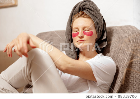 Young woman with casual clothes, towel on head and eye patches sitting on couch and resting. Relax after taking shower. Cosmetic for women, skin care. Self care, time for yourself. Relaxing at home. 89319700