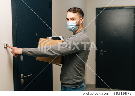 Delivery man in medical mask holding cardboard box and contactless payment POS wireless terminal for card paying, ringing doorbell of customer apartment, looking at camera. Concept of online shopping. Delivery man in medical mask holding cardboard box and contactless payment POS wireless terminal for card paying, ringing doorbell of customer apartment, looking at camera. Concept of online shopping. 89319975