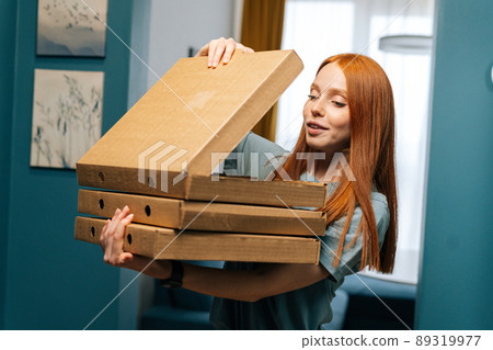 Portrait of happy pretty young woman open box of hot pizza standing at entryway on apartment, looking inside. Hunger female customer received online order from courier with prepared meal for lunch Portrait of happy pretty young woman open box of hot pizza standing at entryway on apartment, looking inside. Hunger female customer received online order from courier with prepared meal for lunch 89319977
