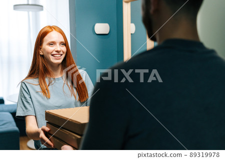 Cheerful young woman receiving paper boxes with hot pizza from unrecognizable delivery man on doorstep at home. Back view of courier male delivering boxes with food to female client at apartment. Cheerful young woman receiving paper boxes with hot pizza from unrecognizable delivery man on doorstep at home. Back view of courier male delivering boxes with food to female client at apartment. 89319978