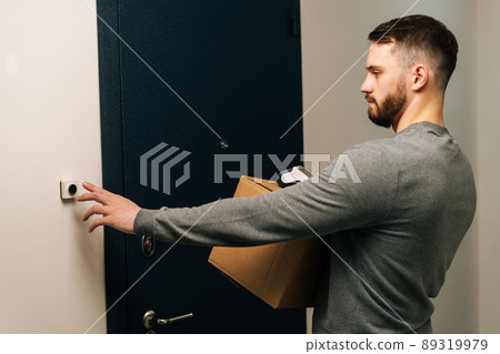 Delivery man holding cardboard box and contactless payment POS wireless terminal for card paying and ringing doorbell of customer apartment. Concept of online shopping. 89319979