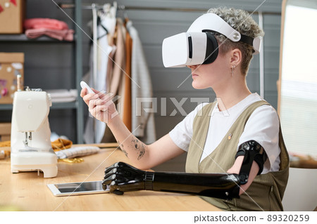 Young female designer or tailor with partial arm in vr headset using remote control while working over new creative project 89320259