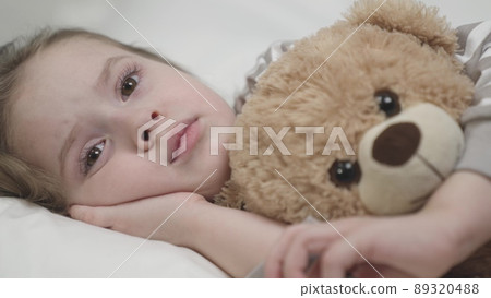 Sleepy kid girl lies bed soft pillow hugging bear toy. Sleepy melancholic small girl lies bed soft pillow hugging chest teddy bear toy. Tired child puts palm under remembering past day with sadness. 89320488