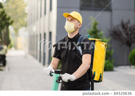 Young caucasian delivery man riding electric scooter in face mask with yellow thermal box backpack. Young caucasian delivery man riding electric scooter in face mask with yellow thermal box backpack. 89320636