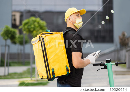 Side view of delivery man in face mask using smartphone and riding push scooter in the city. 89320654