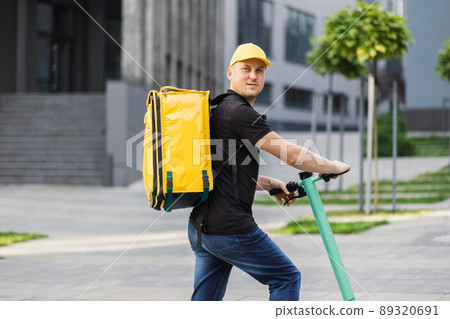 Happy delivery man in yellow cap and thermal insulated bag riding electric scooter on city street. Happy delivery man in yellow cap and thermal insulated bag riding electric scooter on city street. 89320691