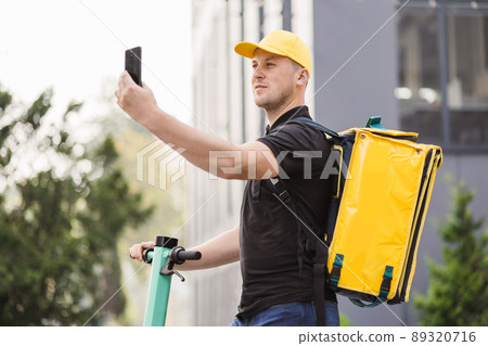 Man courier food delivery with yellow thermal backpack walks street with electric scooter 89320716