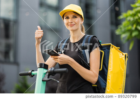 Attractive female courier in yellow cap look at the camera, searching customer on smartphone. Attractive female courier in yellow cap look at the camera, searching customer on smartphone. 89320799