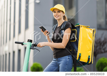 Female courier looking for client address using smartphone riding order on electric scooter. 89320810