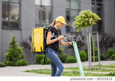 Female courier with thermo backpack browsing address on mobile phone map standing at city street. Female courier with thermo backpack browsing address on mobile phone map standing at city street. 89320818