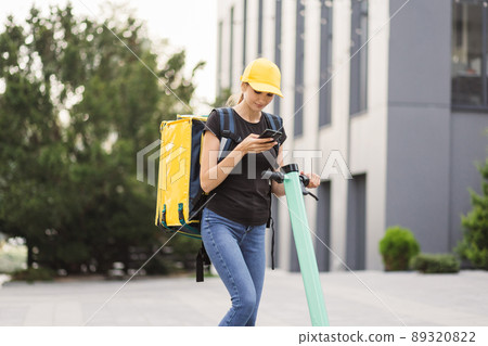 Female courier with thermo backpack browsing address on mobile phone map standing at city street. 89320822
