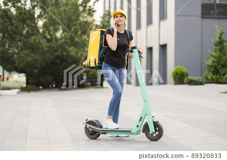 Delivery woman using smartphone while standing in the city with electric push scooter. Delivery woman using smartphone while standing in the city with electric push scooter. 89320833