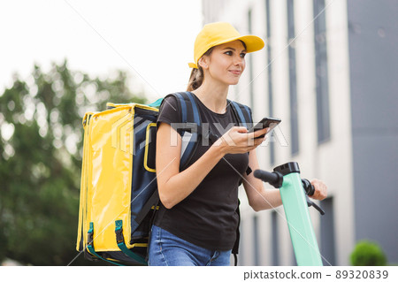 Side view of delivery woman taking order on cellphone standing near scooter on urban playground Side view of delivery woman taking order on cellphone standing near scooter on urban playground 89320839