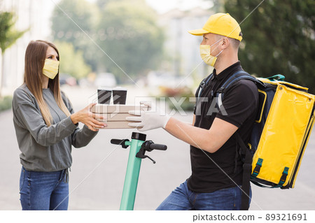 Courier delivering pizza boxes and two cups of coffee to caucasian girl customer in face mask. Courier delivering pizza boxes and two cups of coffee to caucasian girl customer in face mask. 89321691